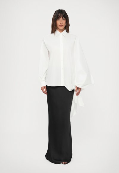 Mujer vistiendo una blusa blanca asimétrica con botones y mangas oversized, y una falda larga negra, de pie sobre un fondo blanco liso.