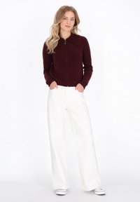 Bordeauxfarbener, gestrickter Zip-Up-Pullover mit großem Kragen, kombiniert mit weiten weißen Hosen und grauen Sneakers, vor einem neutralen Hintergrund.