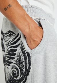 Gray sweatpants with a decorative black design on the upper side, featuring a side pocket and elastic waistband. Soft cotton blend material.