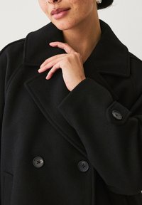 Schwarzer doppelreihiger Mantel mit Kragen, aus schwerem Stoff; verfügt über große schwarze Knöpfe und eine glatte Textur.
