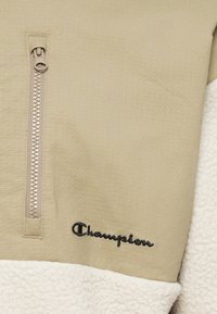 Sudadera de media cremallera en color beige con tejido texturizado en la parte superior, forro polar blanco en la parte inferior y logotipo negro de "Champion" bordado cerca del dobladillo.