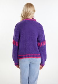 Gruby fioletowy sweter dziany z różowymi paskami akcentującymi, prążkowanym kołnierzem i luźnym krojem. Pokazany z tyłu w połączeniu z jasnoniebieskimi dżinsami.