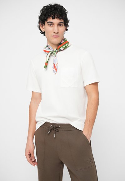 Man with curly hair wears white short-sleeve shirt, brown drawstring pants, and multicolored scarf tied around neck, standing against white background.