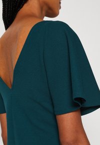 Femme aux cheveux tressés portant une robe vert foncé avec un dos en V profond et des manches courtes évasées.