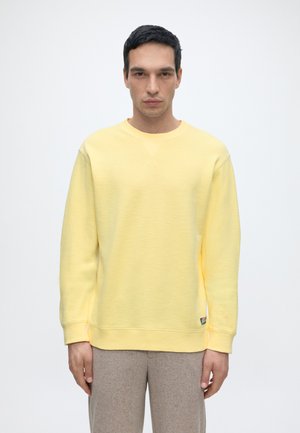 Mann mit dunklen Haaren, der einen gelben Langarm-Sweatshirt und beige Hose trägt und vor einem schlichten hellen Hintergrund steht.