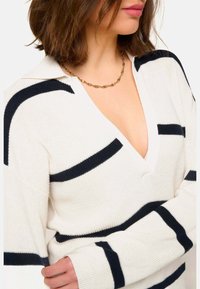 Pull crème avec des rayures navy, design en V, texture côtelée et manches longues. Présente une coupe décontractée et un col plat. Collier en chaîne dorée.