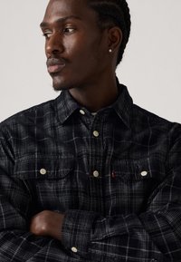 Black and gray checkered flannel shirt with two chest pockets and white buttons; fabric appears soft with a slight texture. Red logo tag visible.