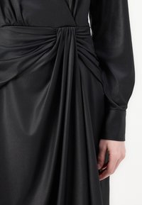 Juoda suknelė, pasižyminti minkšta, blizgia medžiaga, su drapiruotomis detalėmis ties juosmeniu ir ilgomis rankovėmis. Lygus tekstūra, elegantiška konstrukcija.