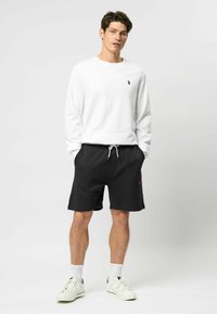 Witte crewneck sweatshirt, zwarte drawstring shorts, eenvoudig ontwerp met rode logo-accent, witte sneakers, gedragen met witte sokken.