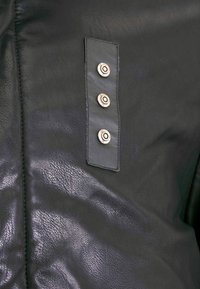 Zwarte leren kleding met een gladde textuur, voorzien van een rechthoekige patch en drie metalen drukknoopjes aan één kant.