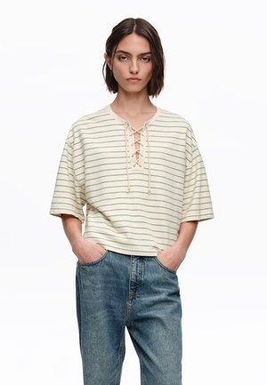 Kobieta o krótkich, ciemnych włosach, ubrana w kremowo-zielony top na sznurowanie w paski oraz niebieskie jeansy, z rękami w kieszeniach, na białym tle.
