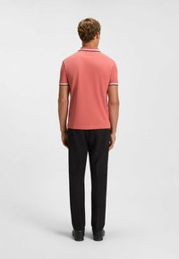 Rosa polo-skjorta med kontrasterande svart och vit randig krage och manschetter, i kombination med svarta byxor och svarta skor, sedd bakifrån.