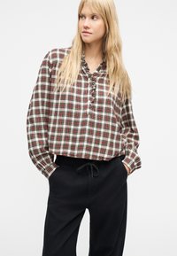 Γυναίκα με μακριά ξανθά μαλλιά, που φοράει ένα λευκό, κόκκινο και μαύρο καρό μπλουζάκι και μαύρο παντελόνι, με τα χέρια στις τσέπες, κοιτάζοντας προς τα δεξιά.