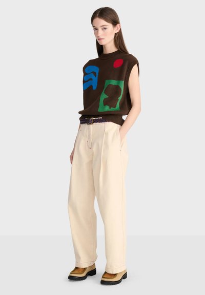 Femme portant un débardeur marron avec des imprimés abstraits colorés, un pantalon large crème, une ceinture multicolore et des chaussures beige avec semelles noires.