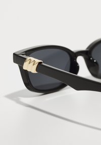 Ochelari de soare negri cu un cadru gros, lentile întunecate și un accent aurie lângă balama, având un design geometric pe braț.