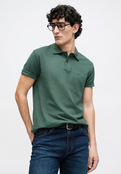 Polo vert avec col et manches retroussées, tissu texturé, petit logo sur la poitrine, porté avec un jean en denim bleu et une ceinture noire.