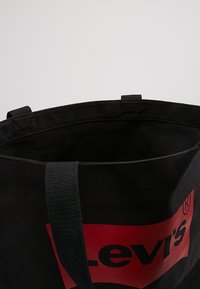 Svart kanvas toteväska med en framträdande röd Levi's-logotyp, öppen upptill med två tygremmar för bärande, inga ytterligare utsmyckningar.