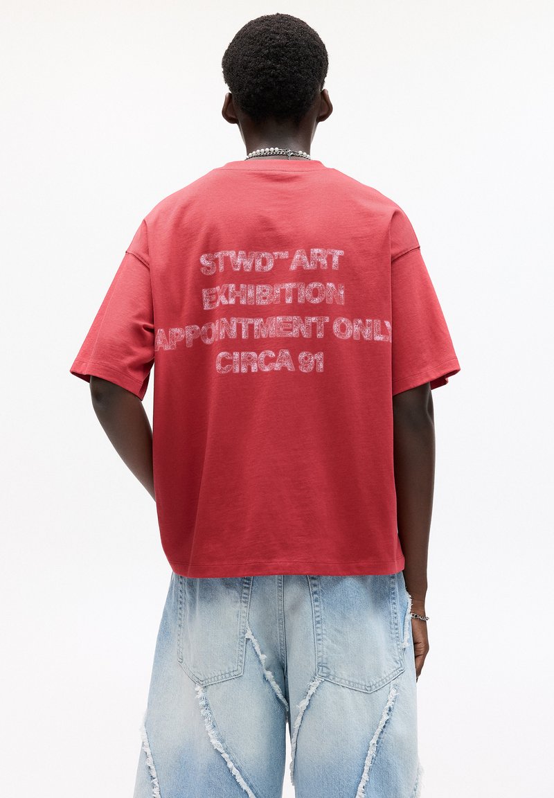 Rød bomulds t-shirt med en løs pasform, der har hvid tekst på ryggen med teksten "STWD’ART UDSMYKNING KUN EFTER AFTALE CIRCA 91."