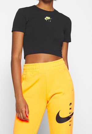 Kobieta w czarnym krótkim t-shircie Nike Air i jaskrawożółtych spodniach dresowych z wieloma czarnymi logo Nike na lewej udzie.