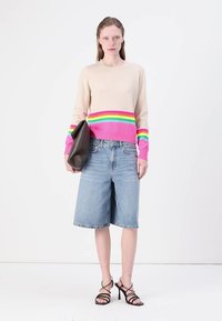 Strikkegenser i beige med rosa, grønn og gul horisontale striper, kombinert med lyseblå denimshorts og svarte sandaler med stropper.