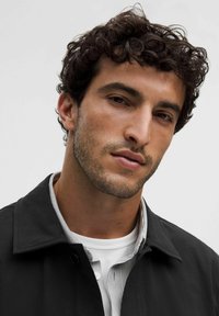 Joven con cabello rizado oscuro y ligera barba que lleva una chaqueta negra sobre una camisa blanca y una camisa de botones a rayas.