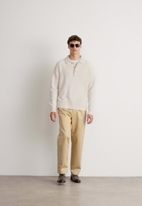 Man met donkere zonnebril, crèmekleurige gebreide trui, beige broek en bruine schoenen die tegen een effen lichte muur staat.
