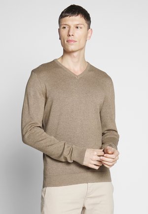 Brązowy sweter w serek wykonany z lekkiego materiału, z długimi rękawami oraz ściągaczami przy mankietach i dole. Noszony z jasnobeżowymi spodniami.