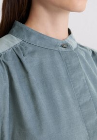 Helles blaues Hemd mit Kragen und gefalteten Schultern, das mit einem einzelnen Knopfdetail und glattem, strukturiertem Stoff ausgestattet ist.