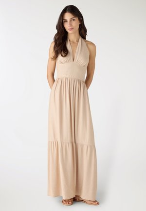 Donna con lunghi capelli scuri che indossa un abito lungo beige senza maniche con collo halter e sandali bassi, in piedi davanti a uno sfondo bianco.