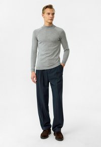 Hellgrauer, taillierter Pullover mit hohem Kragen; gerippte Bündchen und Saum. Kombiniert mit dunklen, locker sitzenden Hosen und braunen Schuhen.