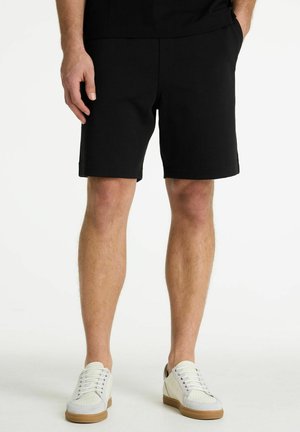 Moški, oblečen v črne kratke hlače do kolen in bele superge z gumijastimi podplati, stoji na enobarvnem belem ozadju.