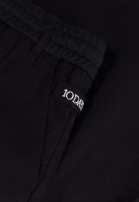 Zwarte joggingsbroek met een elastische tailleband, zijzak en een wit label logo "10DA" gestikt op de zak. Zachte, gestructureerde stof.