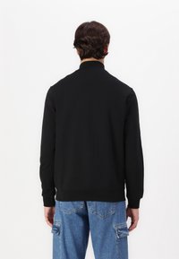 Sudadera negra con cuello simulado, puños y dobladillo acanalados. La parte trasera presenta una textura suave, combinada con unos jeans cargo de color azul claro.