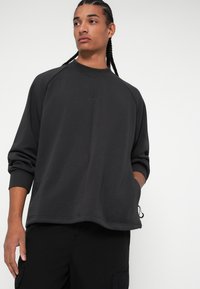 Tumši pelēks sporta krekls ar brīvu piegriezumu, ribotām piedurknēm un mazu logotipu centrā. Izgatavots no mīksta, teksturēta auduma. Ikdienišķs dizains.