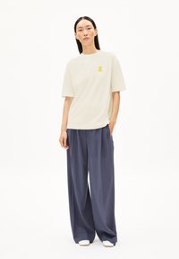 Lichtbeige T-shirt met korte mouwen en een gele graphic, gecombineerd met marineblauwe wijde broek. De outfit heeft een relaxed fit en een zachte textuur.