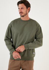 Sudadera verde hecha de una tela suave, con cuello redondo, hombros caídos y una pequeña etiqueta con logo en el dobladillo inferior.