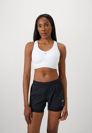 Femme athlétique debout, portant un soutien-gorge de sport blanc et un short de course noir en couches, contre un fond léger uni.
