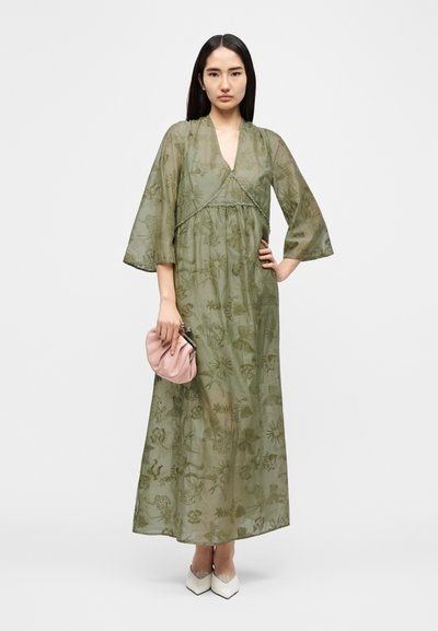 Μακρύ φόρεμα χακί-πράσινο με μανίκια καμπάνα και περίτεχνη βοτανική κεντητική, σε συνδυασμό με ροζ τσαντάκι χειρός και λευκά παπούτσια μυτερά.