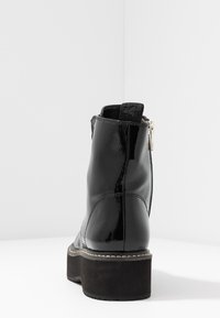 Botte de cheville noire brillante avec semelle épaisse noire, fermeture éclair à l'arrière, détails cousus, et languette en tissu au talon, photographiée sur fond blanc.