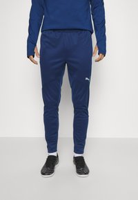 Marineblauwe sportbroek met taps toelopende benen, gemaakt van een gladde stof. Kenmerkt zich door een lichtblauwe zijstrip en een klein wit logo aan de linkerkant.