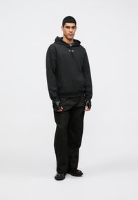 Moletom preto com bolso frontal e capuz, combinado com calças largas escuras. Apresenta luvas sem dedos e sapatos pretos brilhantes.