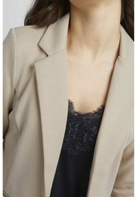 Béžový blazer s texturovanou tkaninou, širokými klopami a přizpůsobeným střihem, vrstvený přes černý top se krajkovým lemem.
