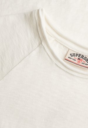 Hellcremefarbenes Shirt mit kurzen Ärmeln und geripptem Rundhalsausschnitt. Das Baumwollmaterial hat eine dezente Struktur; das Etikett trägt die Aufschrift "Superdry."