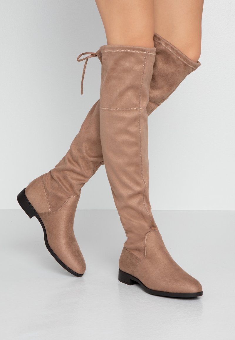 Bottes plates taupe montant au-dessus du genou, bouts ronds, détails cousus, petits talons noirs et fermeture par lien à l'arrière, portées jambes croisées.