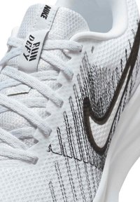 Chaussure de sport blanche avec une tige en mesh texturé, des rayures noires et un logo swoosh proéminent. Elle est équipée de lacets plats et d'une semelle rembourrée.