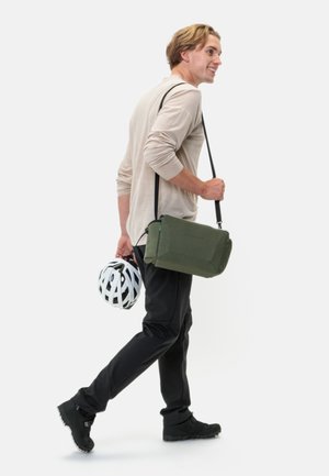 Borsa a tracolla verde con un design testurizzato, cinturino regolabile e chiusura con zip, abbinata a un casco da bicicletta bianco e pantaloni neri.