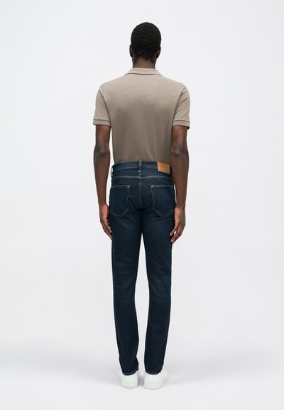 Sötétkék farmernadrág, testhezálló szabással és kontrasztos varrással, bézs pólóval és fehér tornacipővel párosítva.