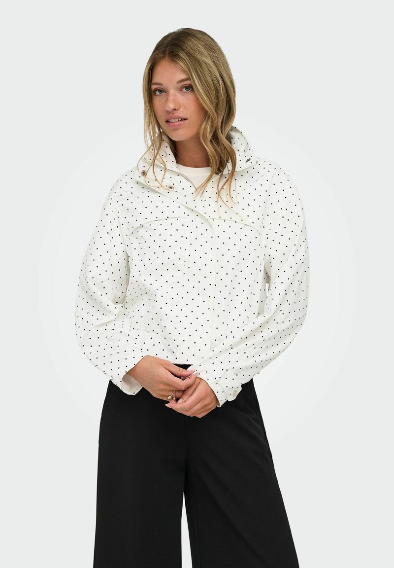 Femme aux cheveux blonds portant une veste blanche à pois et un pantalon noir, debout devant un fond clair uni.