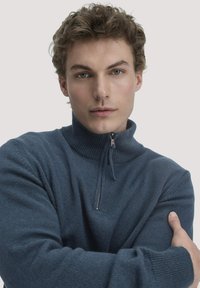 Blauer Reißverschluss-Pullover mit hohem Kragen, gerippter Textur und langen Ärmeln. Verfügt über einen metallischen Reißverschluss. Das Modell hat die Arme verschränkt und trägt ihn.