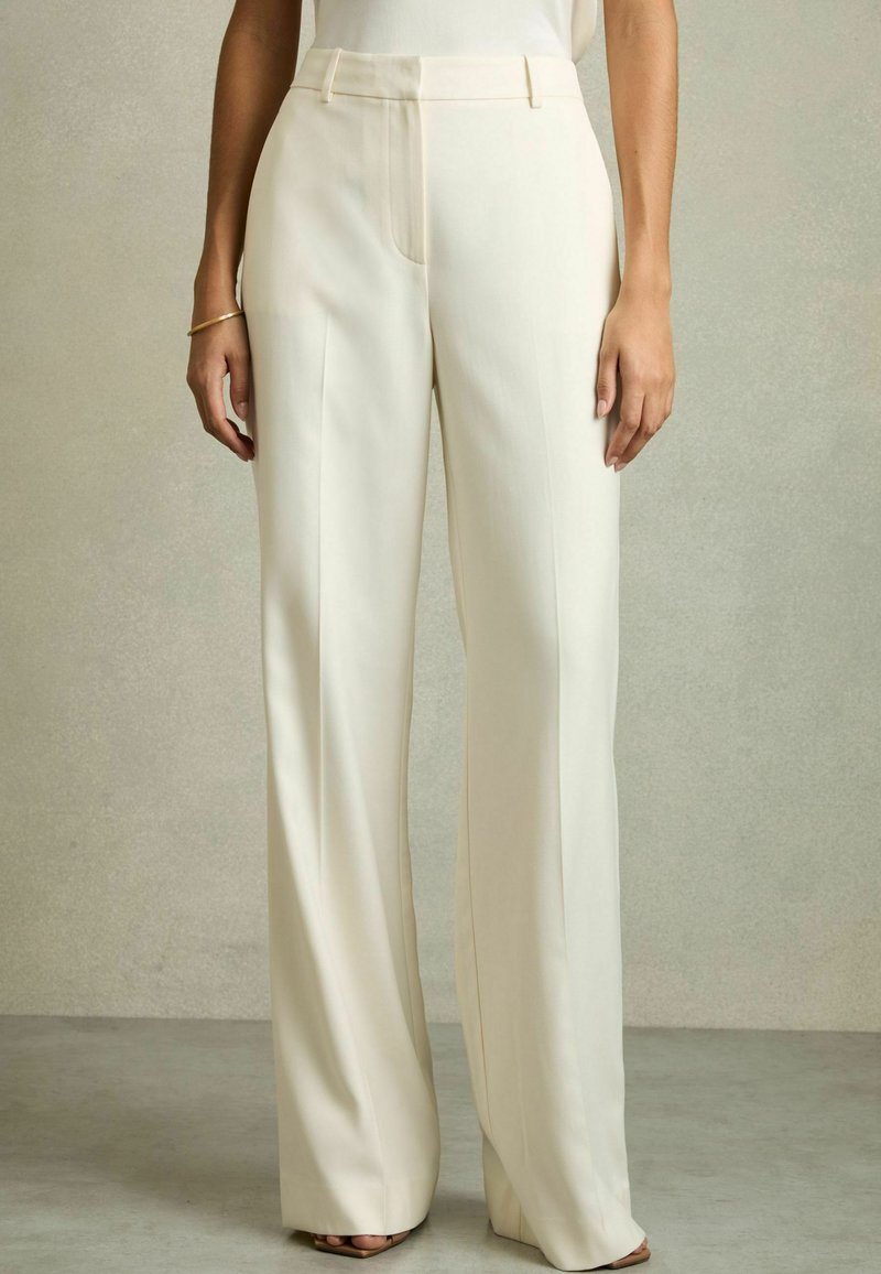 Kalhoty s širokými nohavicemi v off-white látce, s hladkým povrchem, předními záhyby a přizpůsobeným střihem, v kombinaci s jednoduchým topem a minimální obuví.